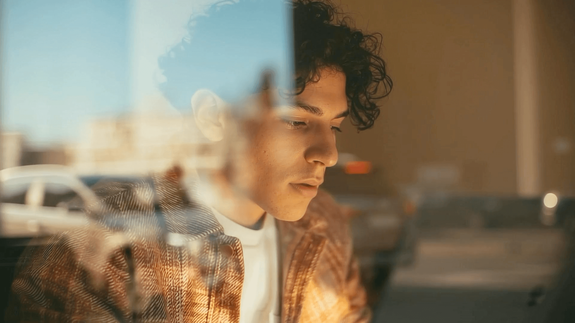 Joven de cabello rizado mirando hacia abajo con calma, detrás de una ventana